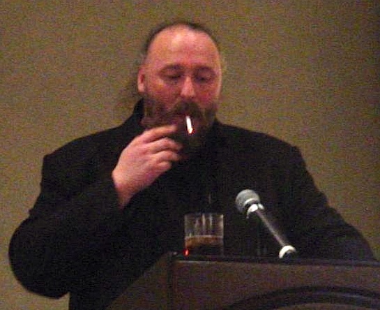 Warren Ellis smoking a cigarette and drinking whiskey at a podium